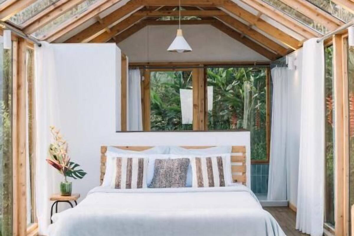 A cozy bedroom with a wooden bed, a hanging light fixture, and large windows offering a view of greenery outside.