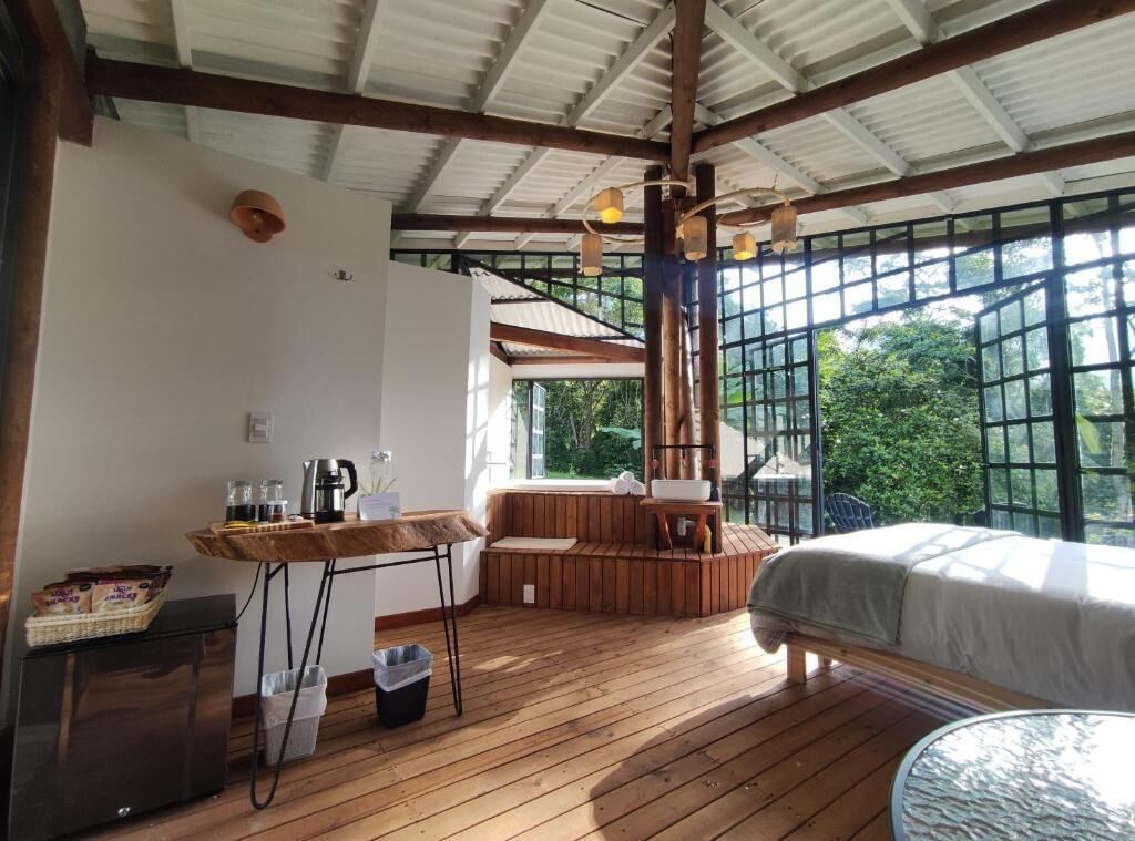 Une pièce chaleureuse avec un plancher en bois, un lit, une petite table avec une bouilloire et une grande fenêtre donnant sur un paysage verdoyant.