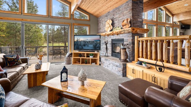A spacious living room with a stone fireplace, wooden ceiling, and large windows overlooking a deck. - Golf-View | 6K SqFt, 7 BR/8 Ba, 6 Kings | SHARC Pass | Bikes | Pool Table + Bar (Sunriver)