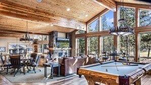 A spacious living room with a pool table, a fireplace, and large windows offering a view of the outdoors. - Golf-View | 6K SqFt, 7 BR/8 Ba, 6 Kings | SHARC Pass | Bikes | Pool Table + Bar (Sunriver)