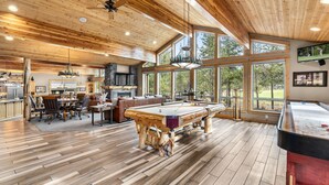 A spacious indoor area with a pool table, wooden flooring, and large windows offering views of the outdoors. - Golf-View | 6K SqFt, 7 BR/8 Ba, 6 Kings | SHARC Pass | Bikes | Pool Table + Bar (Sunriver)
