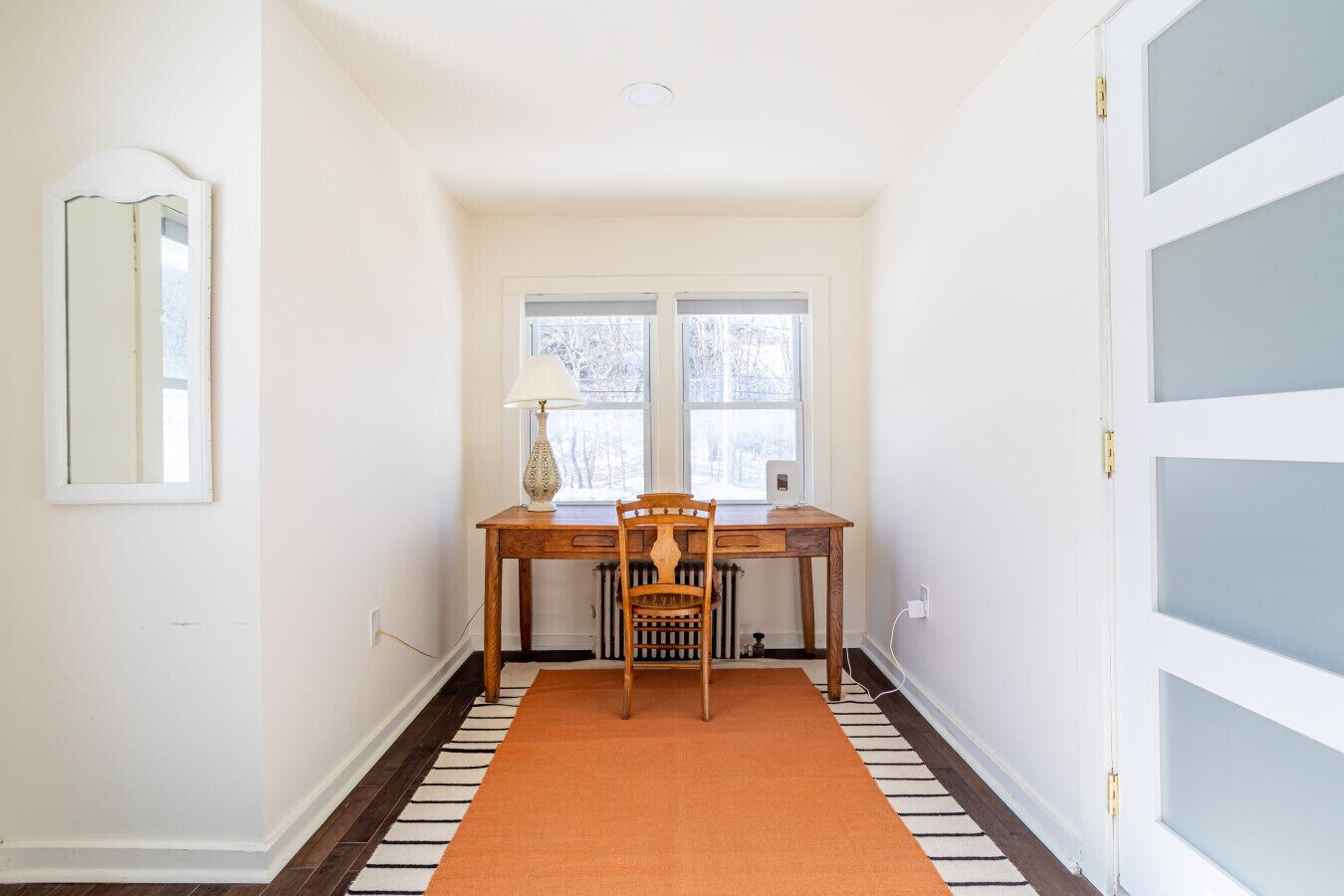 A hallway with a wooden table and chair, a window with a view, and a striped rug.