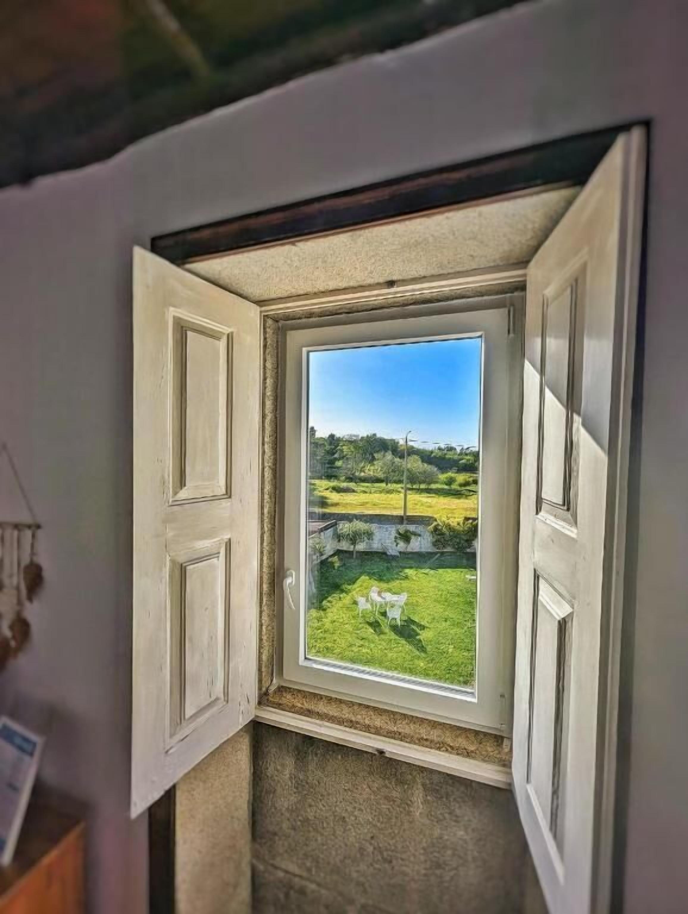 Una habitación con ventana que da a un espacio exterior con césped, una valla blanca y algunas sillas blancas.
