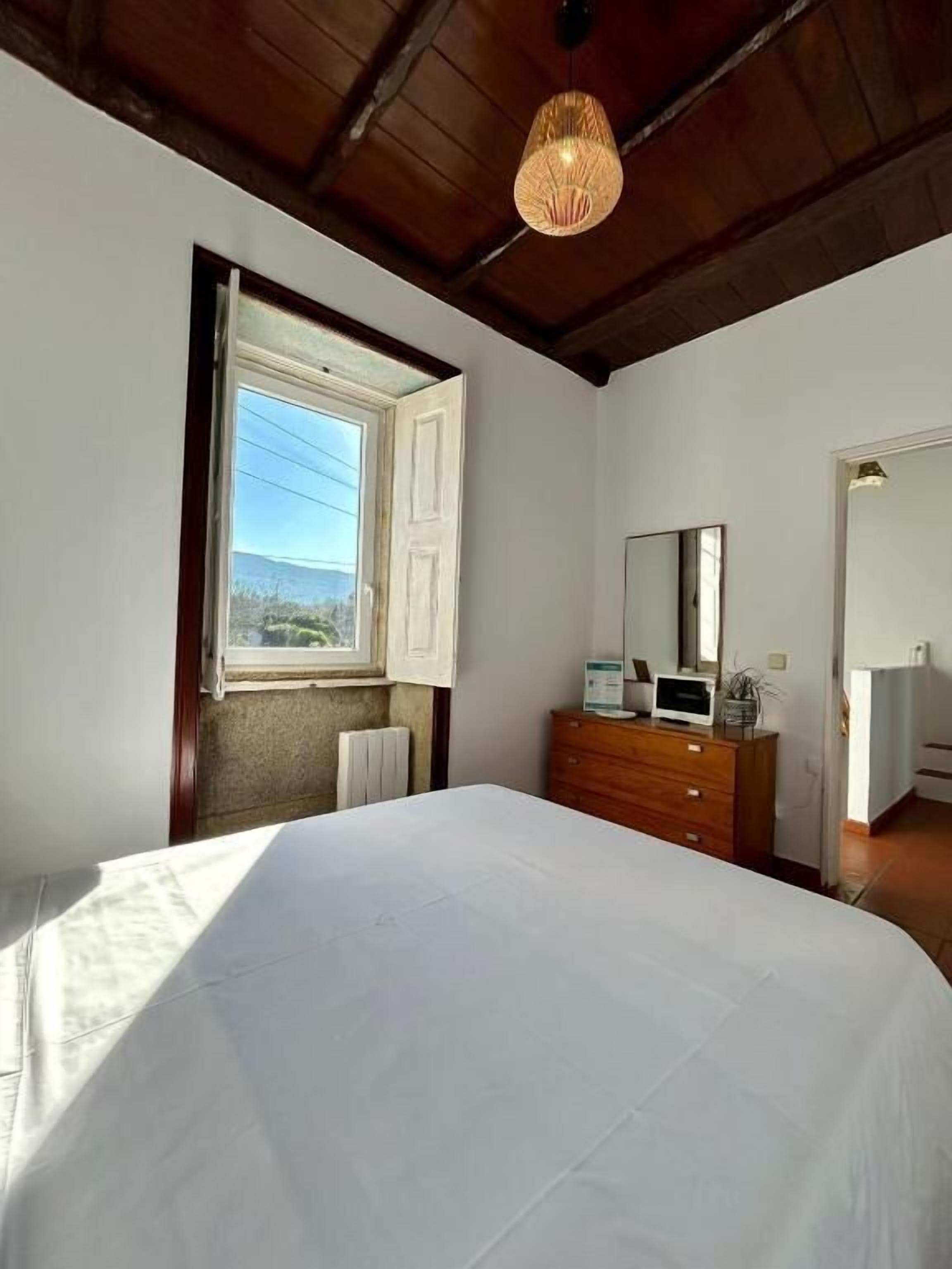 Un dormitorio con una cama grande, un aparador de madera, un espejo y una ventana con un persiano abierto.