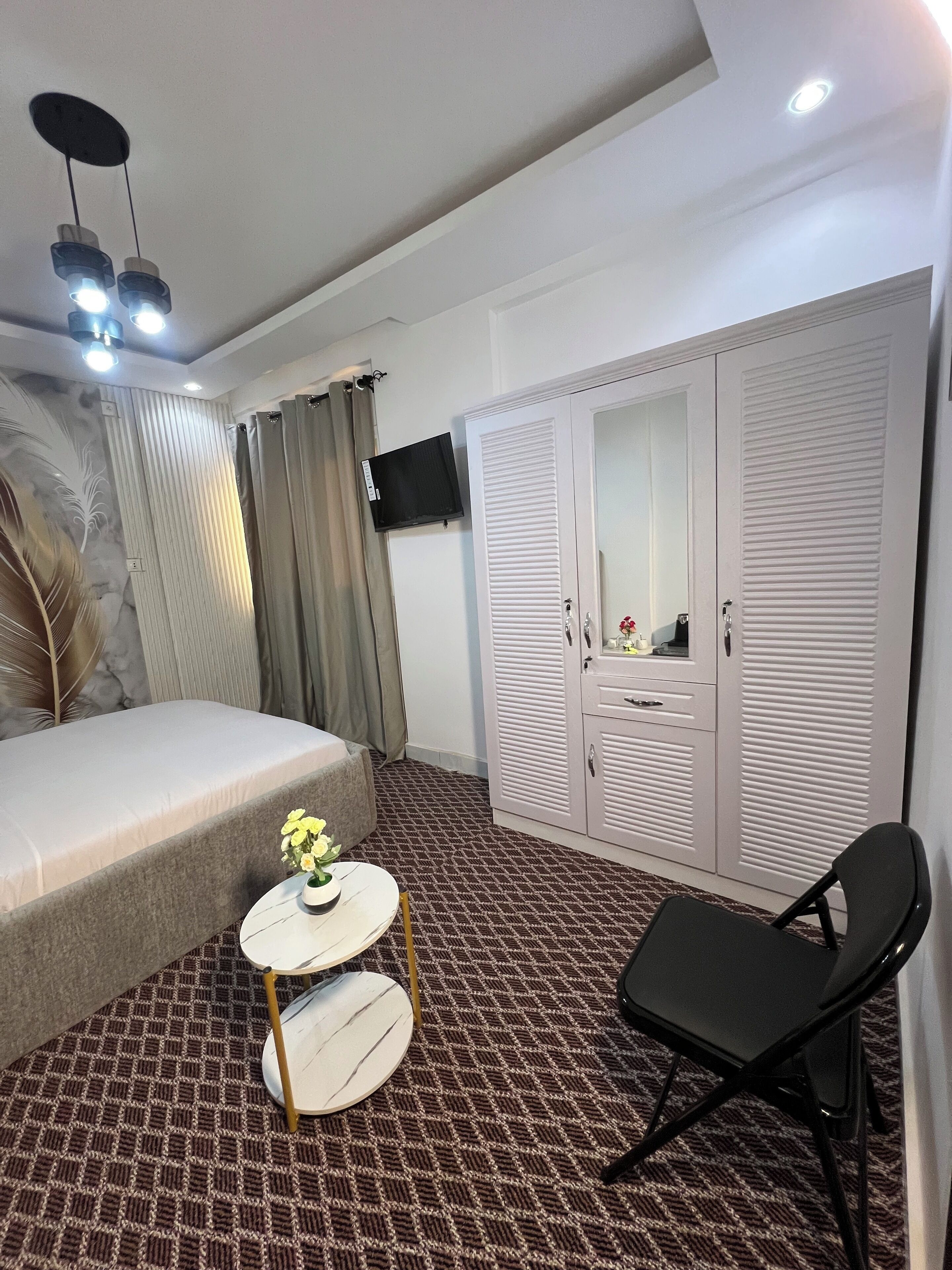 Une chambre à coucher avec un lit, une petite table ronde ornée d’un vase de fleurs, une chaise noire, une armoire avec un miroir et un téléviseur fixé au mur.