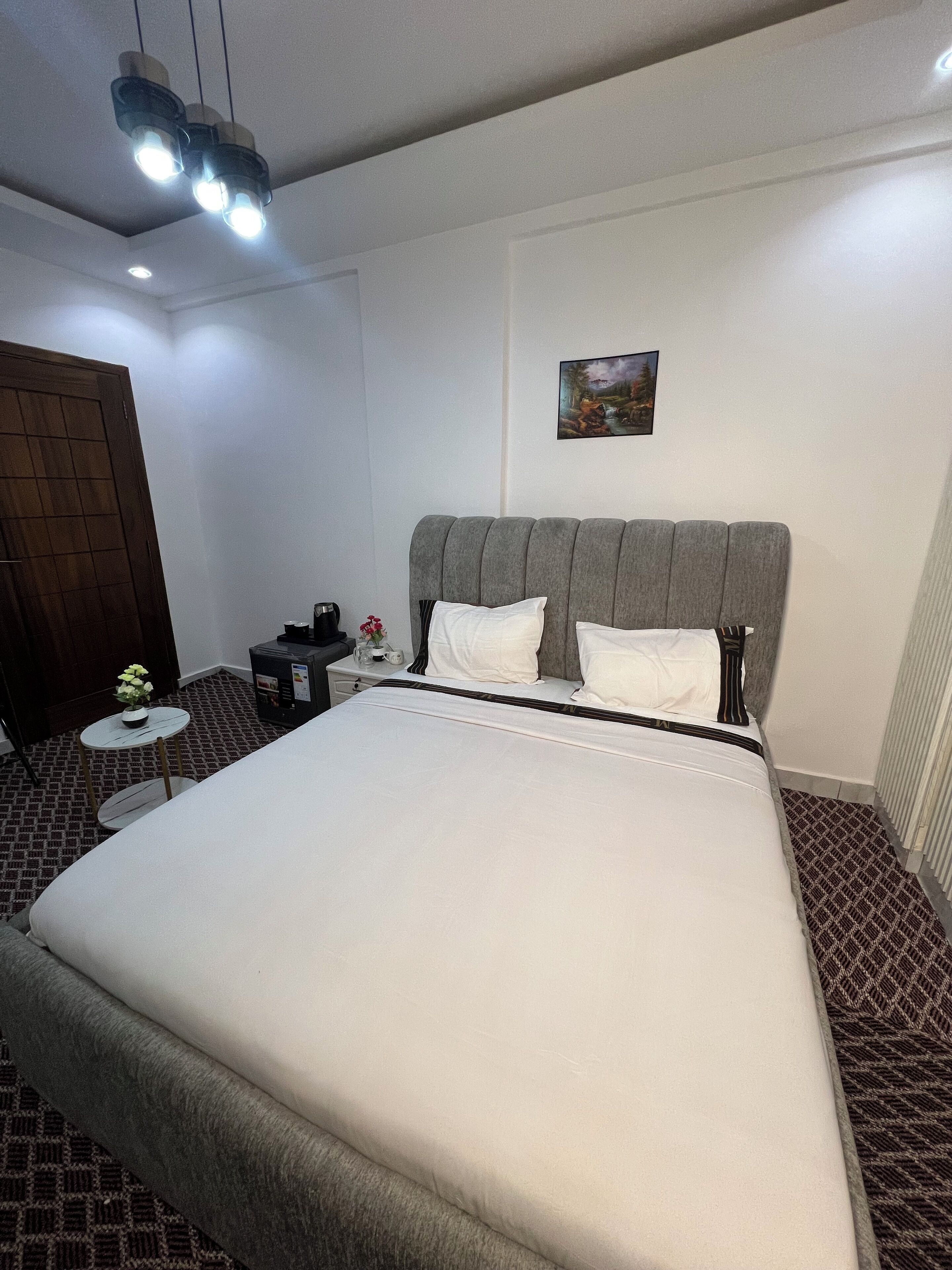A neatly arranged bedroom with a large bed, a small round table with a vase, a nightstand, and a painting on the wall.