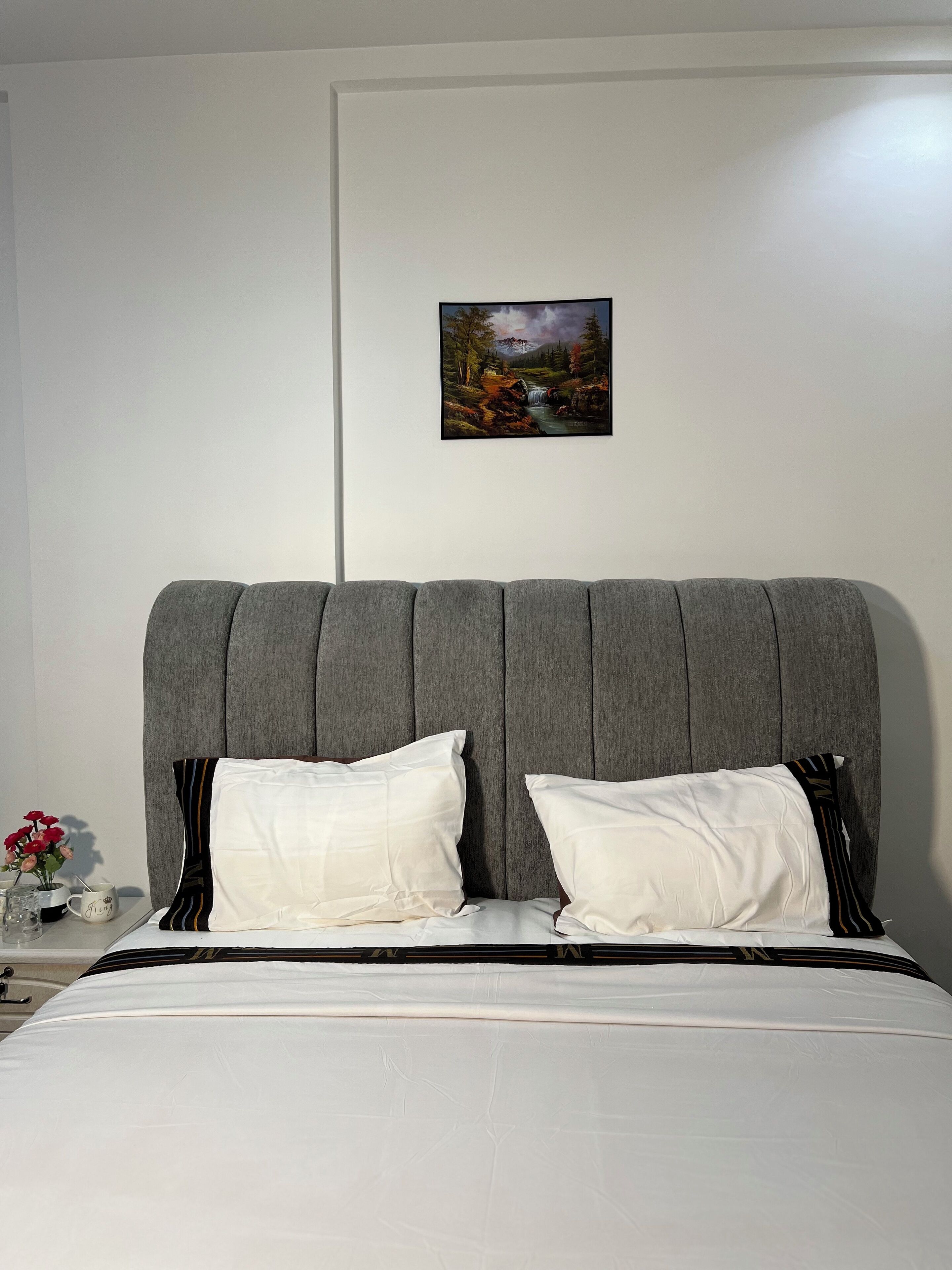 A bed with a grey upholstered headboard, white bedding, and two white pillows. A framed landscape painting hangs on the wall above the bed.