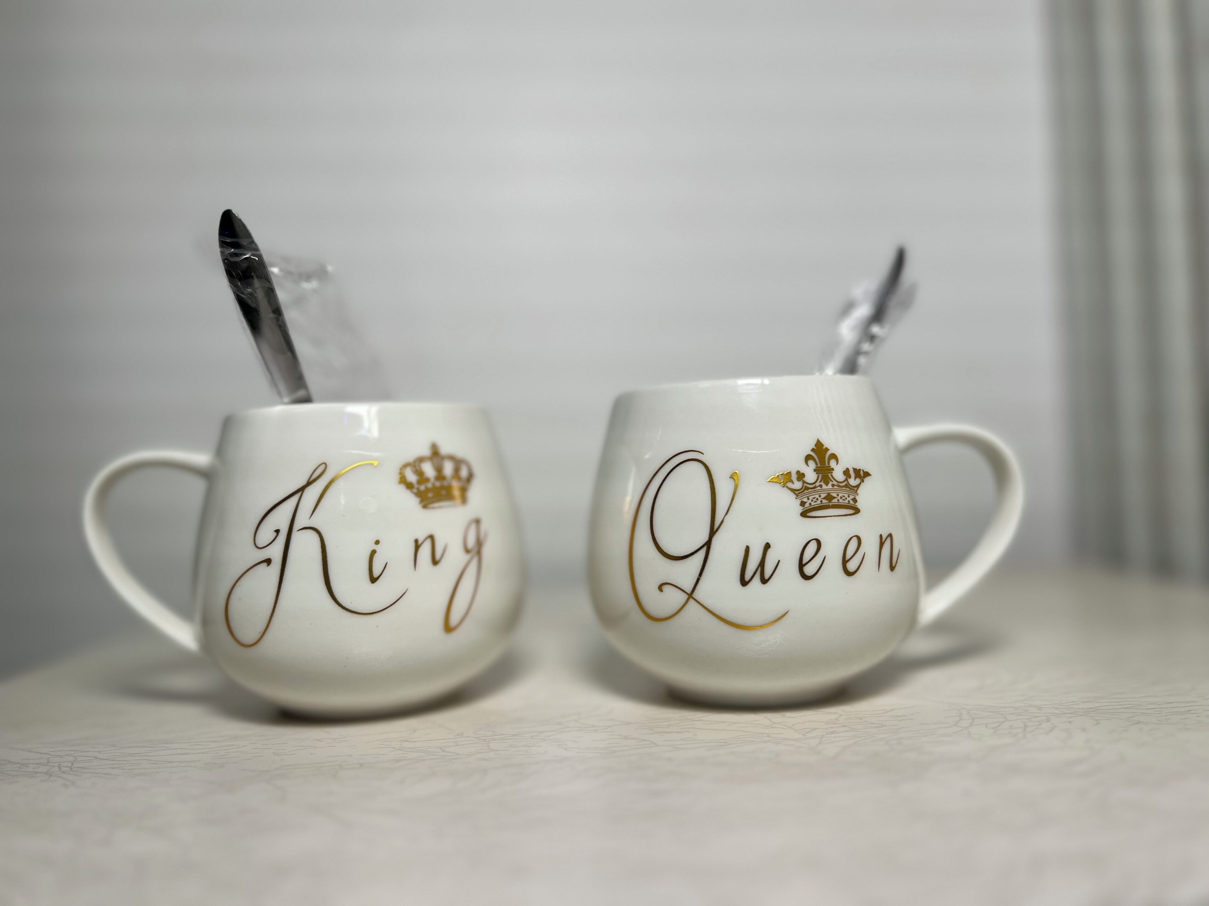 Twee witte keramische mokken met gouden letters en kroonmotieven, waarop 'King' en 'Queen' staan.