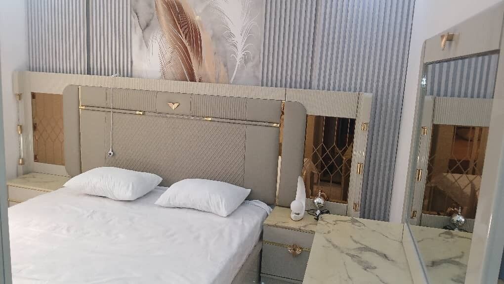 Un dormitorio con cama, mesitas de noche, un espejo y un panel decorativo en la pared.