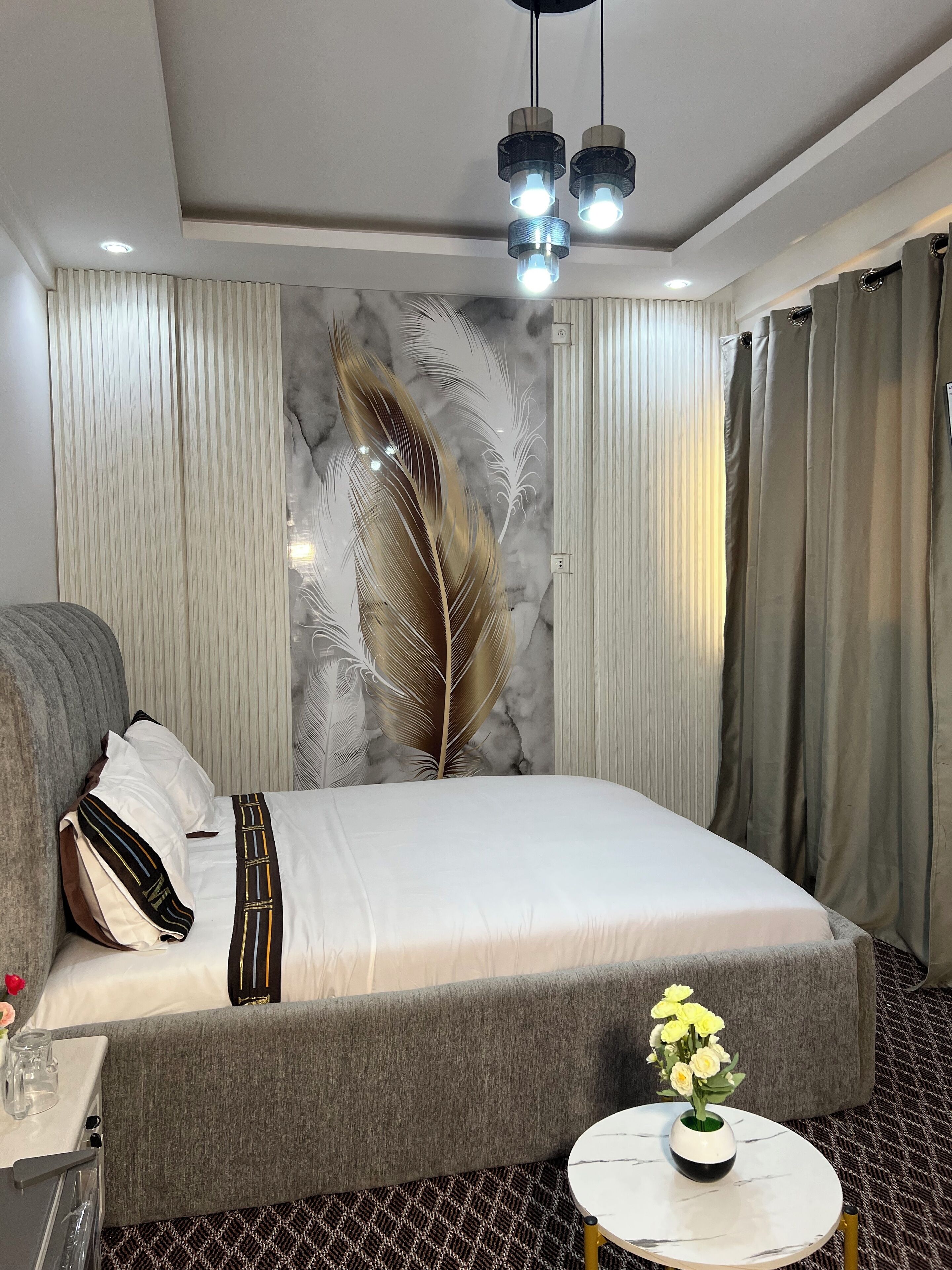 Una habitación de hotel moderna con una cama grande, cortinas grises, un arte de pared decorativo con plumas y una pequeña mesa redonda con un jarrón de flores.