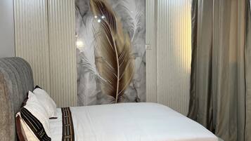 Una habitación de hotel moderna con una cama grande, cortinas grises, un arte de pared decorativo con plumas y una pequeña mesa redonda con un jarrón de flores.