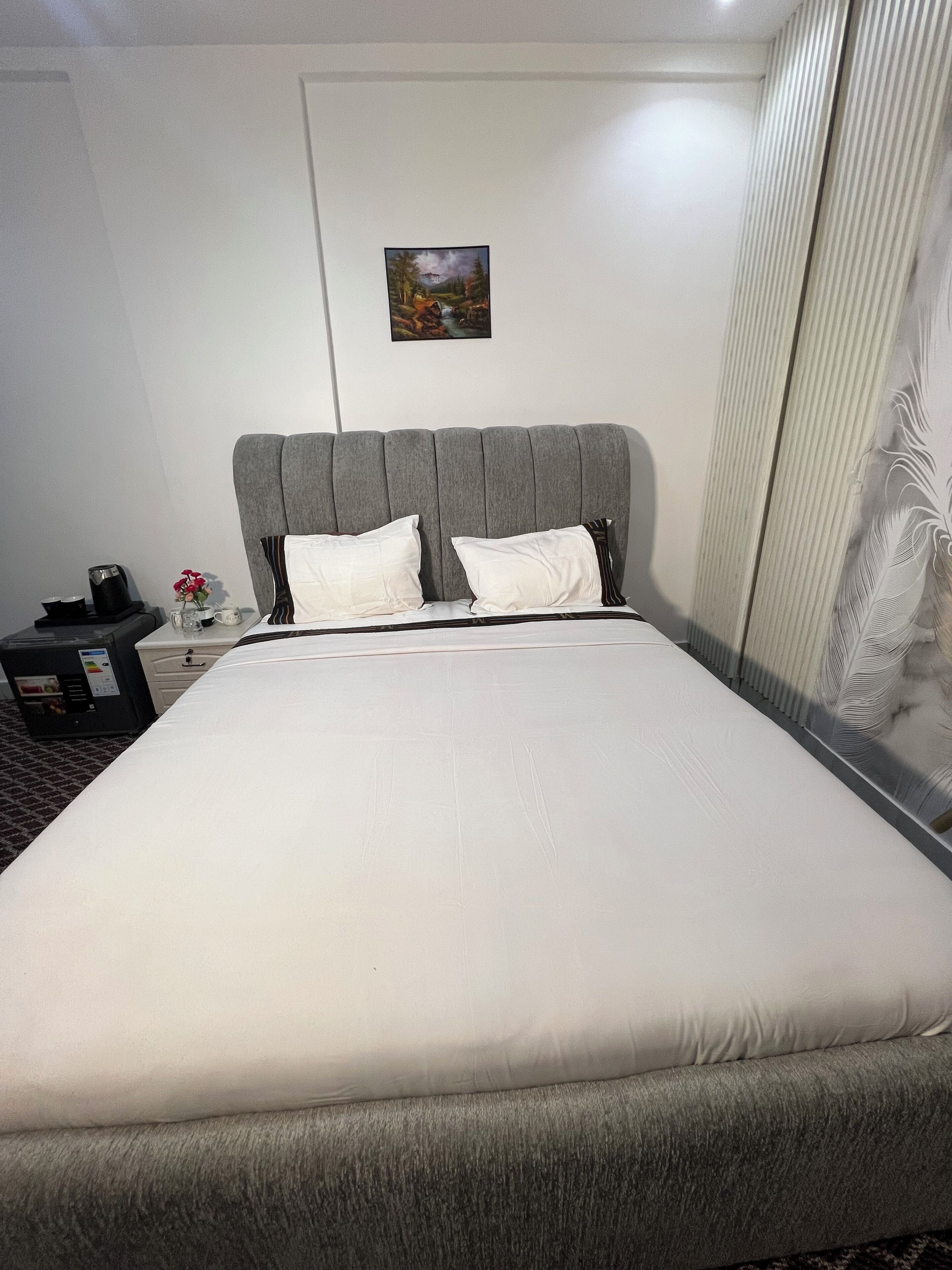 Un letto rifatto con precisione, con una testiera grigia e biancheria da letto bianca, un comodino con una lampada e un quadro appeso al muro.