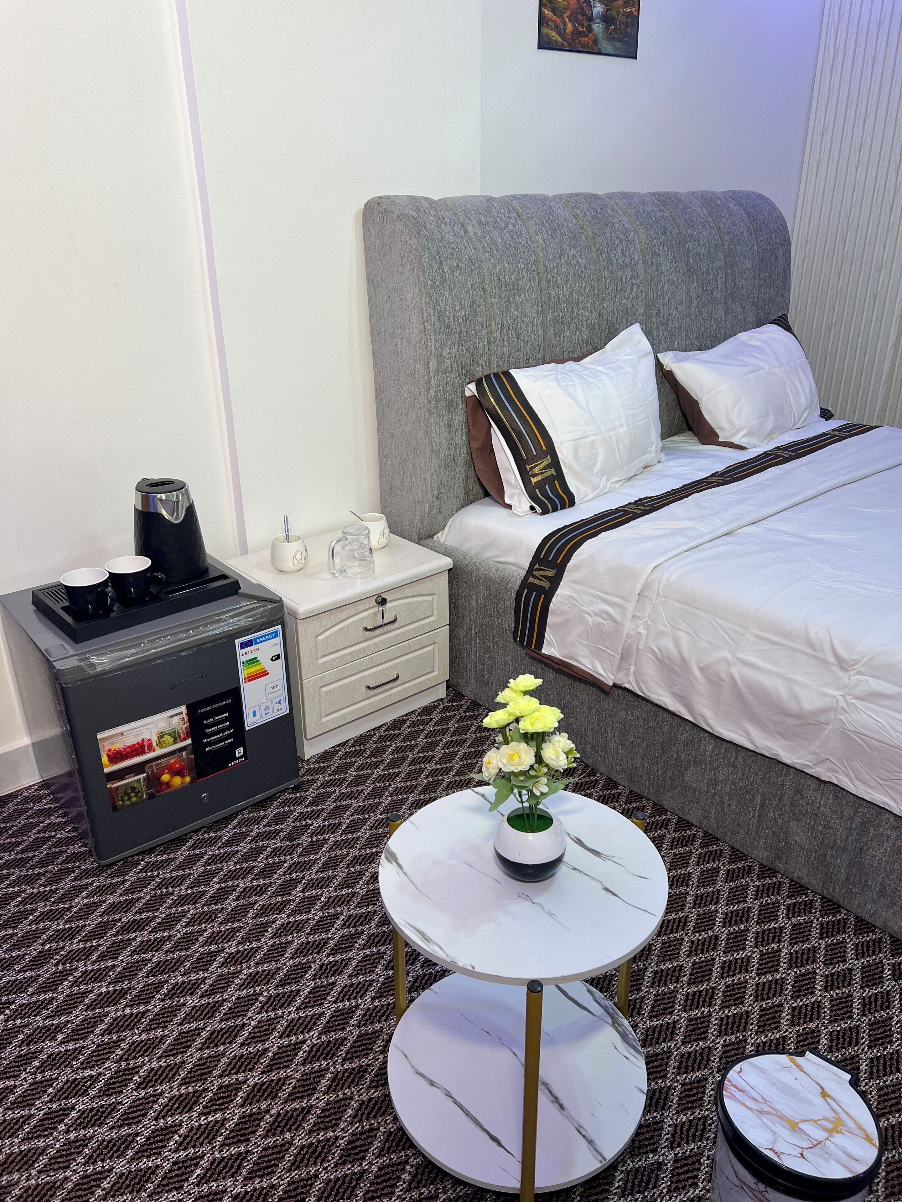 Une chambre d’hôtel comprenant un lit, une table de chevet, une machine à café et une petite table sur laquelle est posé un vase de fleurs.