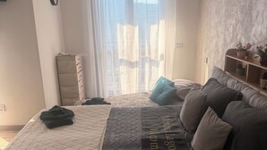 Ein Schlafzimmer mit Bett, Nachttisch, Kommode, einem Fenster mit Vorhängen und einem Wandregal mit Dekoration.