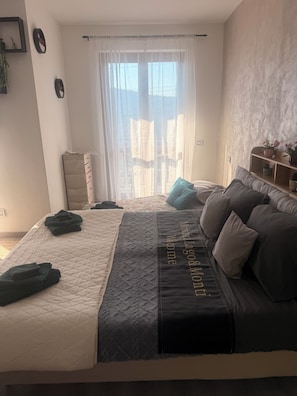 Un dormitorio con cama, mesita de noche, cómoda, ventana con cortinas y estante de pared con objetos decorativos.