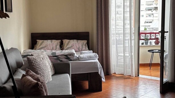 A modern living room with a sofa, a bed with neatly folded towels, a hanging light bulb, and a view of a balcony with plants. - Arena Stadium Apartment (Tiranë)
