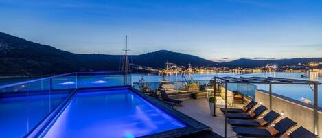 A rooftop infinity pool with lounge chairs, a wooden deck, and a view of a marina and city lights.
