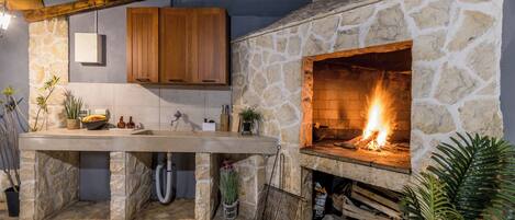 A stone fireplace with a fire, a kitchen counter, wooden cabinets, and potted plants.