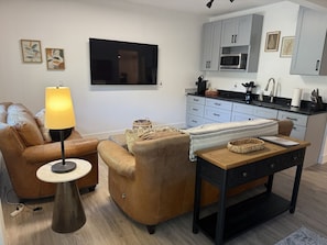 A living room with a sofa, a lamp, a coffee table, and a kitchen area with cabinets and a microwave.