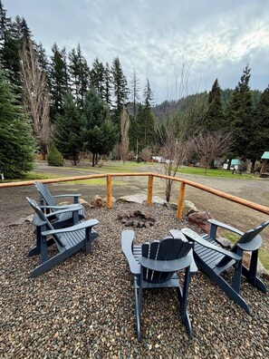 An outdoor seating area with Adirondack chairs and a fire pit, surrounded by a wooden fence and a gravel path.