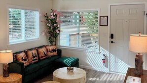 A cozy living room with a green sofa, round wooden ottoman, and a round coffee table. There are two lamps, a door, and a window with blinds.