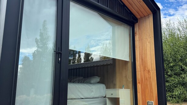 A modern, compact cabin with a large window, a wooden bench outside, and a bed inside. - Hosteria Hainen Cabins, located in El Calafate, 10 minutes from downtown. (El Calafate)