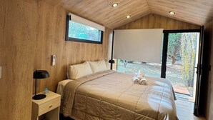 A bedroom with a wooden ceiling, a bed with a beige quilt, a nightstand with a lamp, and a window with a view of the outdoors. - Hosteria Hainen Cabins, located in El Calafate, 10 minutes from downtown. (El Calafate)