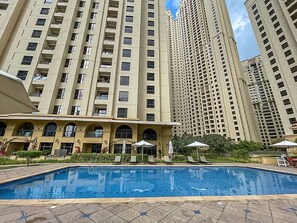 A swimming pool with lounge chairs and umbrellas, surrounded by high-rise residential buildings. - Stayshort - Modern 4 Bed Apartment in JBR That Sleeps 8 (Dubai)