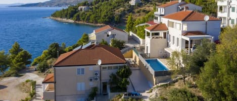 Ett bostadsområde vid kusten med hus som har röda tegeltak, en swimmingpool och utsikt över havet och bergen.