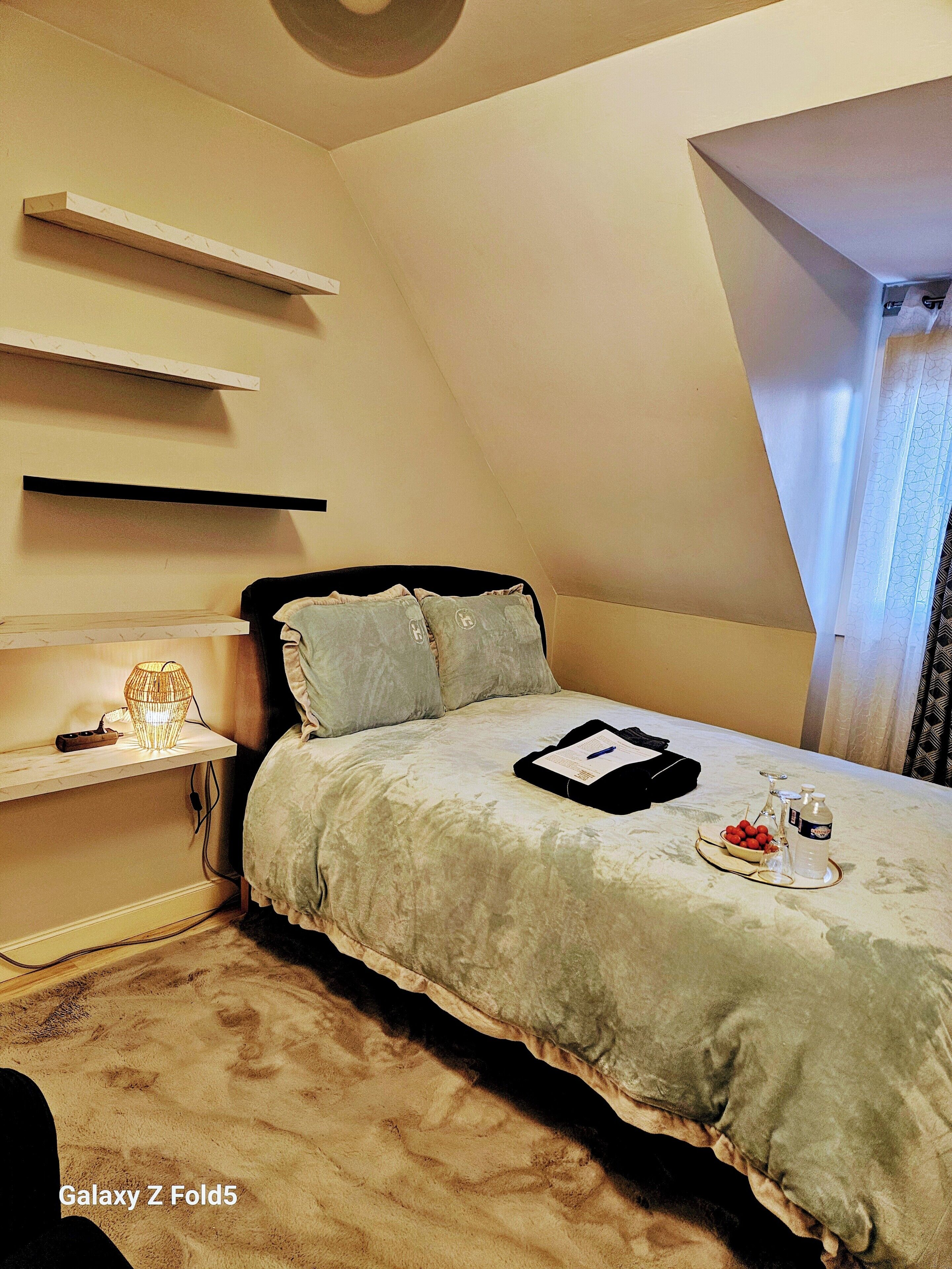 Une chambre à coucher avec un lit, des tables de chevet, une lampe et un plateau sur lequel se trouvent une bouteille et des fruits.