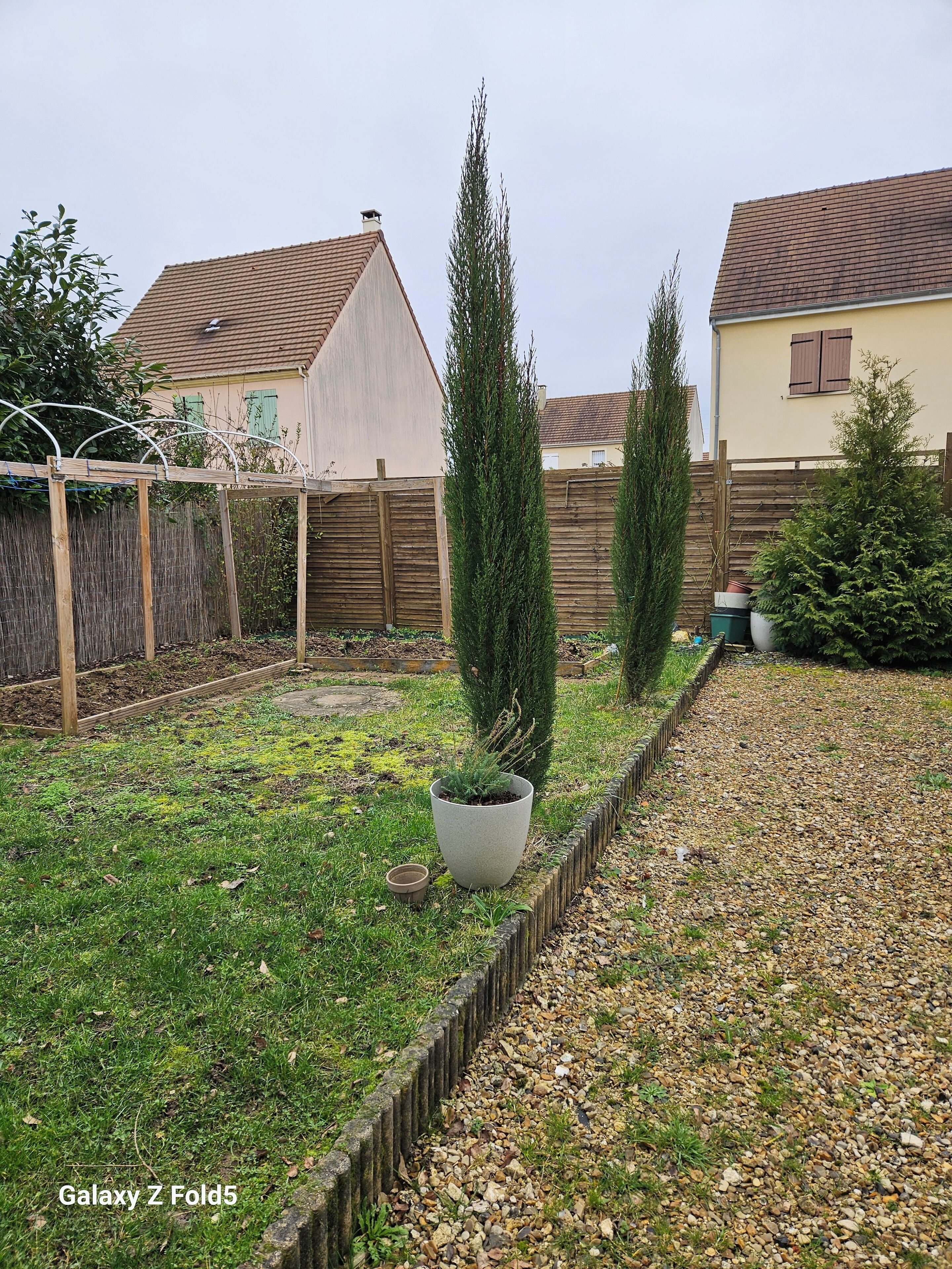 Un jardin arrière d’une maison, avec un parterre de fleurs, un grand arbre à feuilles persistantes et une clôture en bois.