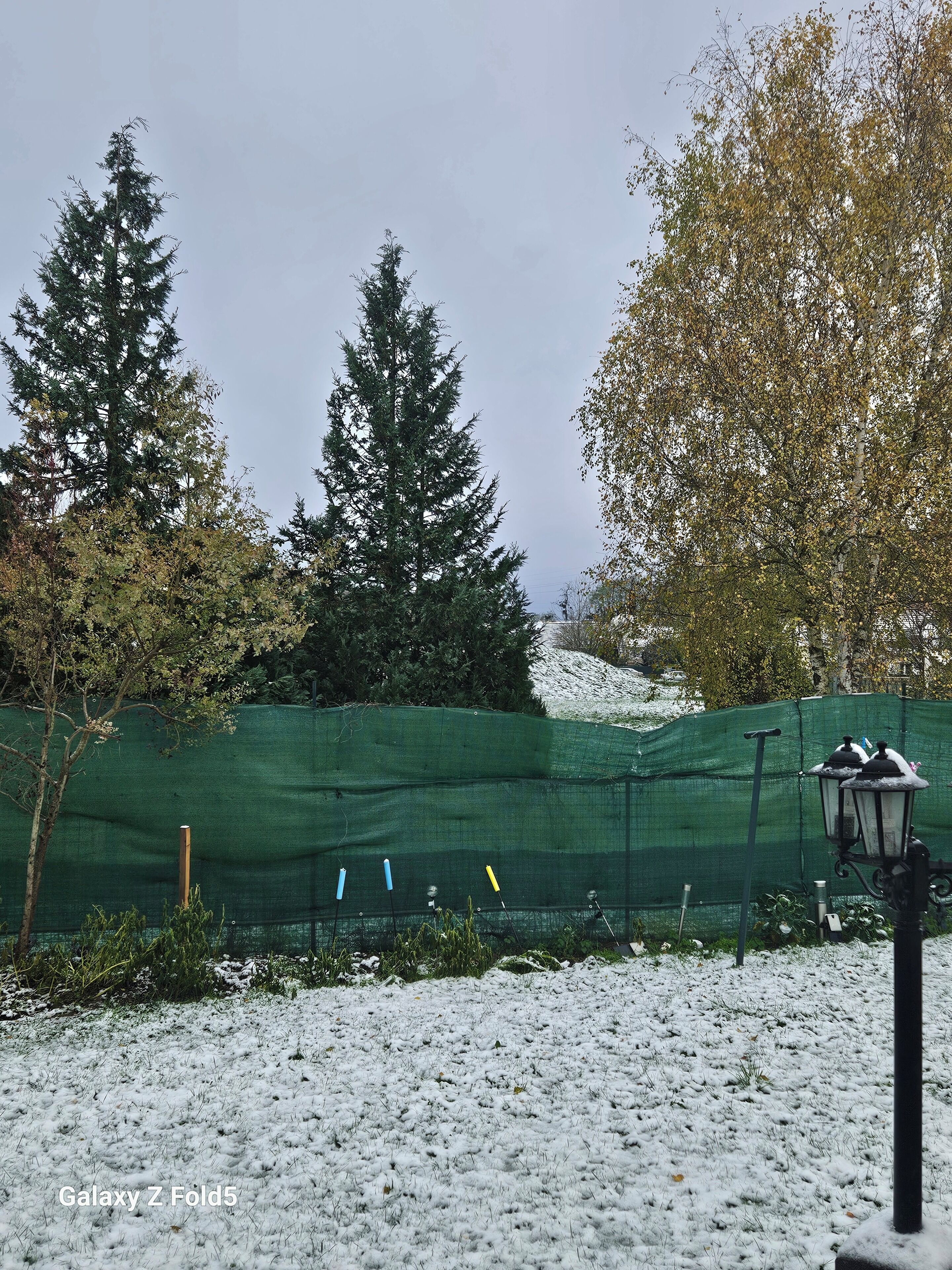 Paisaje nevado con una cerca verde, árboles y una farola.