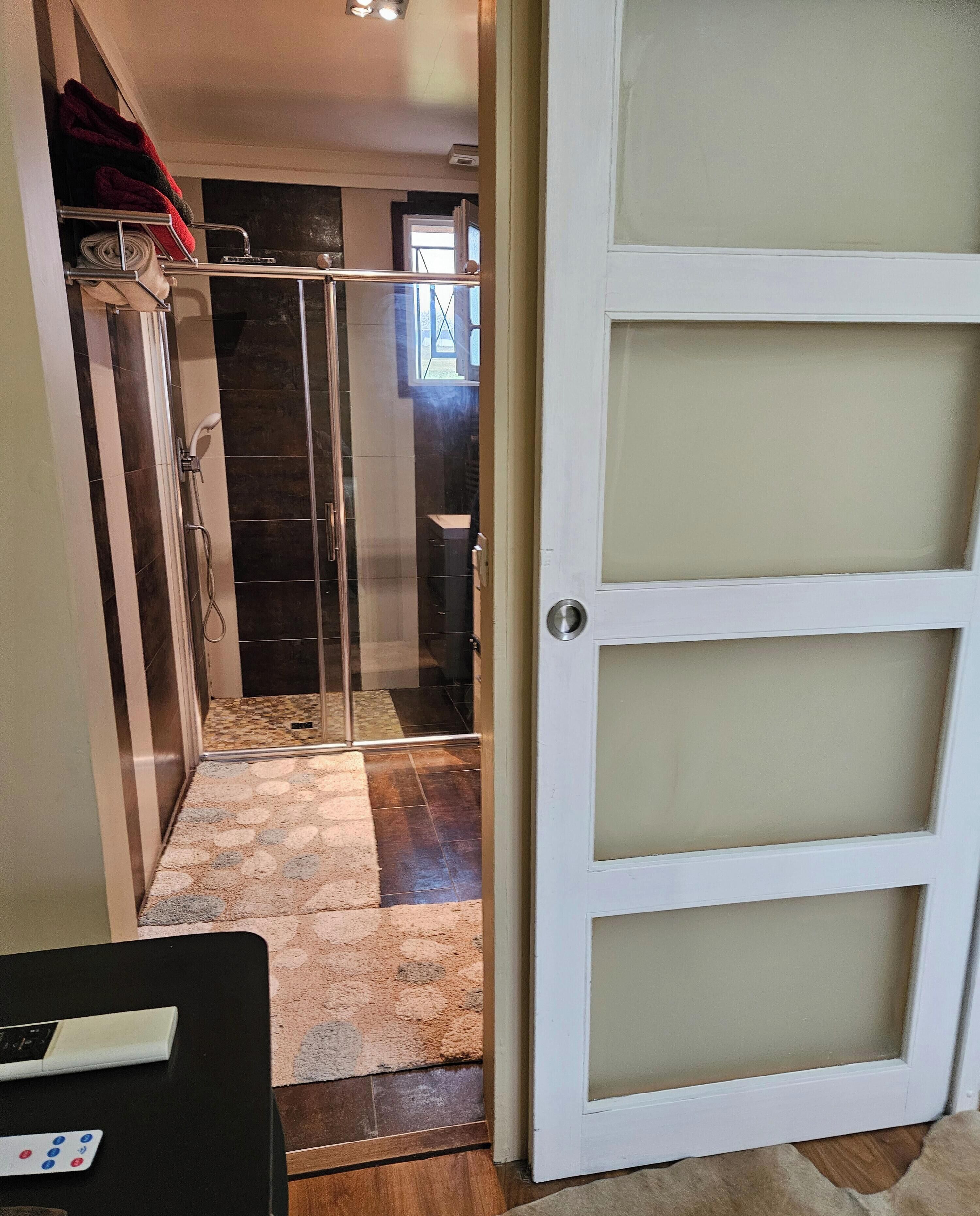 Un baño con cabina de ducha de vidrio, una puerta blanca con paneles esmerilados y un perchero con toallas rojas.
