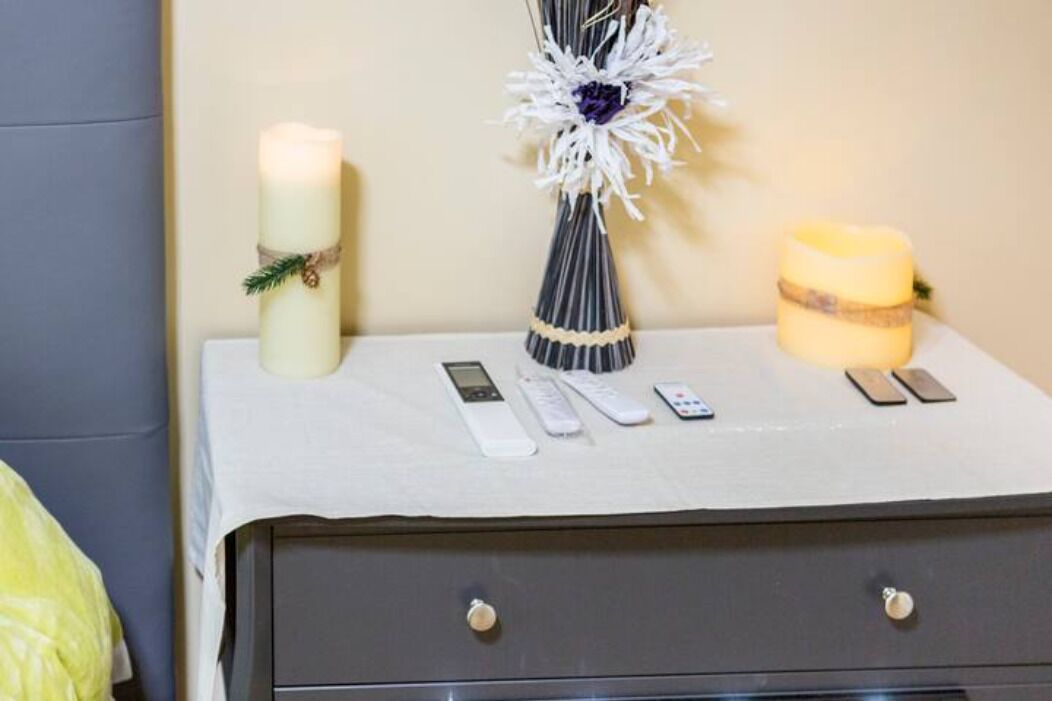 A bedside table with a white cloth, a white candle, a decorative vase, a yellow candle, and several remote controls.
