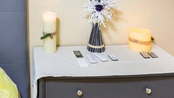 A bedside table with a white cloth, a white candle, a decorative vase, a yellow candle, and several remote controls.