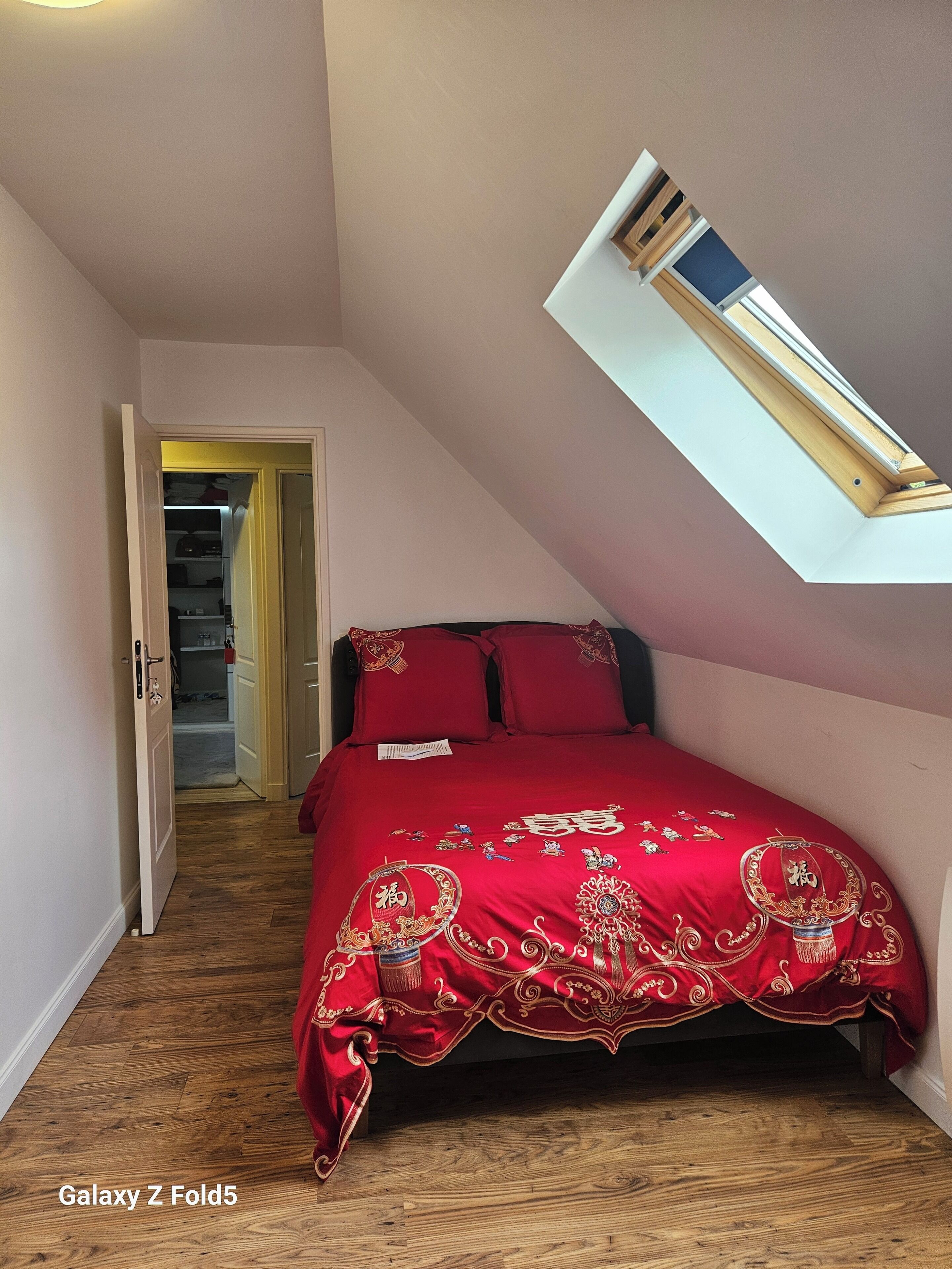 A bedroom with a sloped ceiling, a skylight, a red bedspread, and a wooden floor.