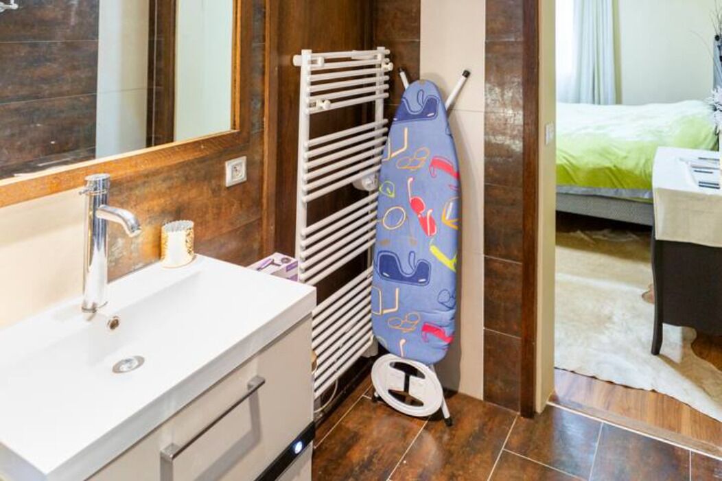 Un bagno con un lavandino bianco, uno specchio, un portasciugamani e un letto visibile attraverso una porta aperta.