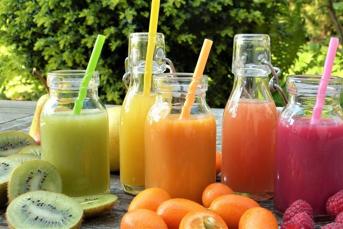 Fire glasflasker med sugerør, hver med en smoothie i forskellige farver, omgivet af frugtskiver.