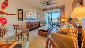 A spacious living room with a large window overlooking the beach, a ceiling fan, and a dining area with a table and chairs. - Sunlit Island Suite w/ Lanai Access & Sunset Ambiance - Experience Live Music! (Hawaii County)
