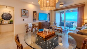 A modern living room with a glass coffee table, wooden chairs, a large flat-screen TV, and a view of the beach through large windows. - Sunlit Island Suite w/ Lanai Access & Sunset Ambiance - Experience Live Music! (Hawaii County)