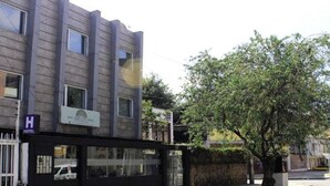 Un bâtiment d’hôtel doté d’une façade moderne, d’une entrée sécurisée par un portail et d’un arbre devant.