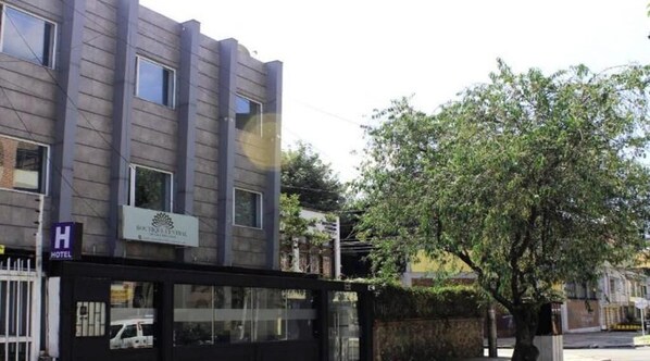 Un bâtiment d’hôtel doté d’une façade moderne, d’une entrée sécurisée par un portail et d’un arbre devant.