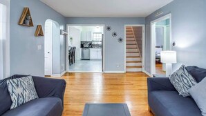 Ein modernes Wohnzimmer mit Holzboden, zwei dunkle Sofas mit gemusterten Kissen, einem kleinen Teppich und einer Treppe, die zu einer weiteren Ebene führt.