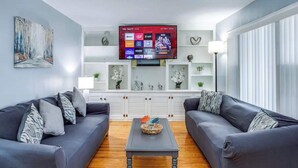 Ein modernes Wohnzimmer mit zwei grauen Sofas, einem Holztisch und einem Wand-Fernseher, der verschiedene Sender zeigt.