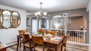 Un coin repas avec une table en bois dressée pour quatre personnes, entourée de chaises. Un lustre est suspendu au-dessus de la table, et la pièce présente un design moderne avec des murs gris et un plancher en bois.