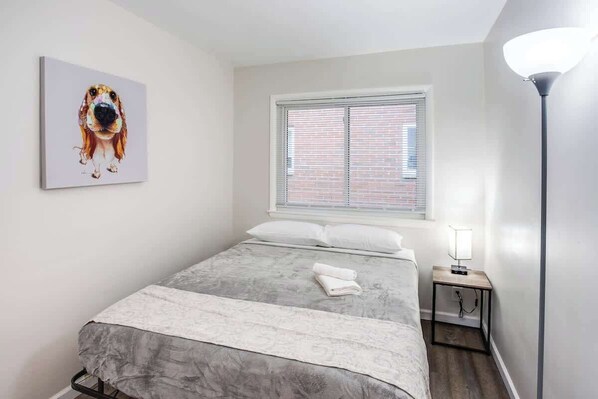 A bedroom with a bed, a window with blinds, a bedside table with a lamp, and a wall-mounted painting of a dog. - 💫Modern & Quiet Suburban Ranch Mins to Dtwn/Stores (East Rochester)