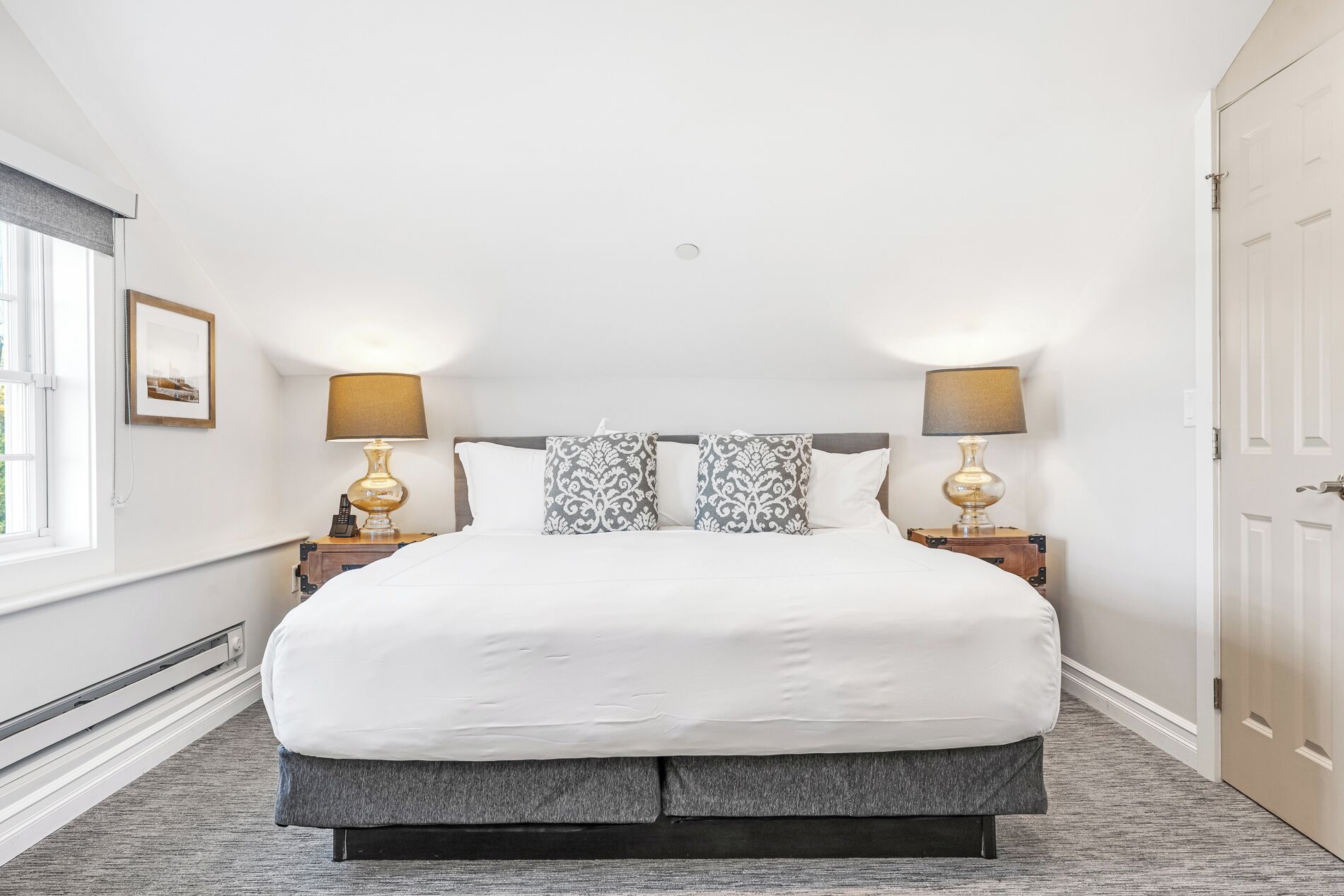 Een netjes opgemaakte slaapkamer met een groot bed, twee bedlampjes, een ingelijst kunstwerk aan de muur, en een deur aan de rechterkant.