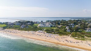 An aerial view of a coastal town with a wide sandy beach, numerous beach umbrellas, and a large building complex with multiple houses. - The Eagle’s Nest Penthouse: 360° Views in the Heart of Watch Hill (Westerly)