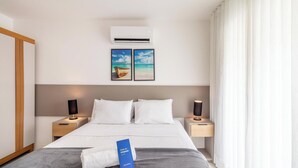 A hotel room with a bed, two bedside tables, a wall-mounted air conditioner, and two framed pictures of a beach scene. - Apt w/ Balcony, BBQ & Pool View Ano0209 (Cabo Frio)