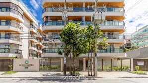 A modern multi-story residential building with balconies, a sign for 'Ocean Garden Condominium Complex', and a tree in the foreground. - Apt w/ Balcony, BBQ & Pool View Ano0209 (Cabo Frio)